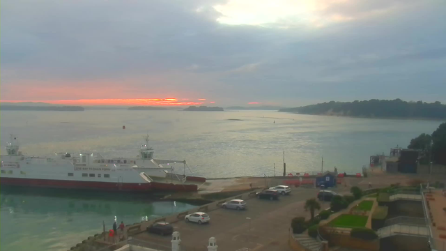 A coastal scene at dawn featuring a calm waterway. In the foreground, a ferry boat is docked at a marina with its bow facing the camera. Several parked vehicles are lined along a pavement near the water. Lush greenery and landscaped areas are visible to the right, leading towards the water. The sky is overcast with hints of orange and pink from a rising sun illuminating the horizon in the distance over calm waters, surrounded by distant hills and shoreline.