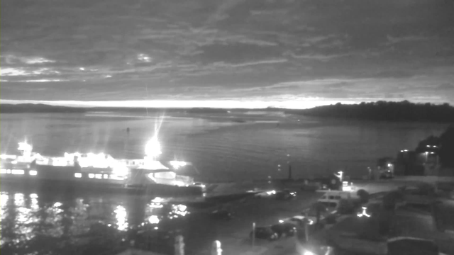 A nighttime scene of a body of water with a faintly illuminated ferry docked at a pier. The water reflects light from the ferry and surrounding buildings. In the background, there are dark silhouettes of land and clouds in the sky, adding depth to the scene, while the horizon is slightly illuminated by a distant light source. There are several vehicles parked along the shore.