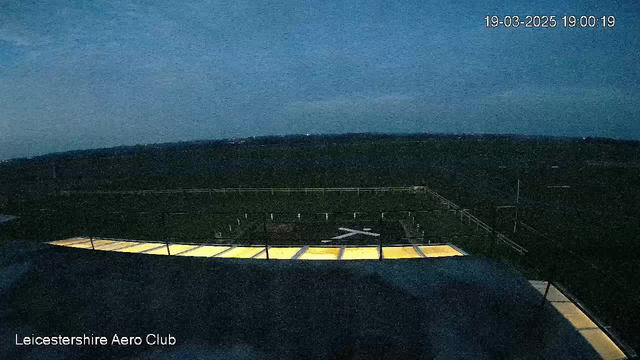 A dark scene showing a flat rooftop with a few yellow light panels along the edge. Below, a grassy field stretches out with several white markers visible in the distance. The sky is overcast and transitioning to twilight, with faint hints of light on the horizon. The time displayed indicates 19:00:19 on March 19, 2025. The lower left corner has the text "Leicestershire Aero Club."