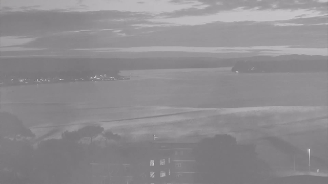 A grayscale image showing a calm body of water extending towards the horizon. The sky is filled with clouds, creating a muted light atmosphere. On the left side, faint lights from buildings or boats are visible along the shoreline, while a distant island is seen on the right side, partially shrouded in mist. The foreground features gentle waves and some indistinct landforms, creating a serene and quiet natural landscape.