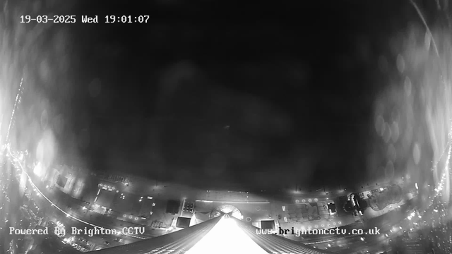 A black and white webcam image showing a cityscape at night. The scene is mostly dark with faint lights visible in the lower part of the image, representing buildings and streetlights. There's a large tower structure in the foreground with a pointed roof leading towards the top of the image. The date and time are displayed in the upper left corner, indicating March 19, 2025, at 19:01:07. The bottom right corner contains the text "Powered By Brighton CCTV" alongside a website URL.