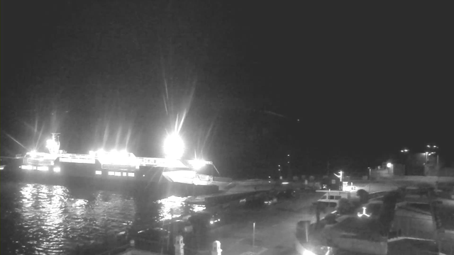 A dark scene at night featuring a large, brightly lit ferry docked at a pier. The reflective water shows shimmering lights from the vessel. In the foreground, there are outlines of vehicles and structures along the dock, with some additional lights visible in the background, indicating other buildings or equipment. The sky is mostly dark, adding to the nighttime atmosphere.