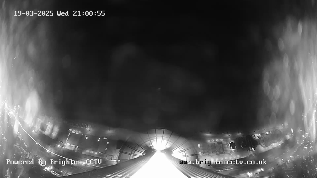 A black and white webcam image showing a city skyline at night. The scene is mostly dark with a bright light source at the center bottom, possibly from a street or building. The layout includes some buildings and streets visible in the darkness, along with faint reflections and highlights from the light. The timestamp and website information are displayed in the corners.