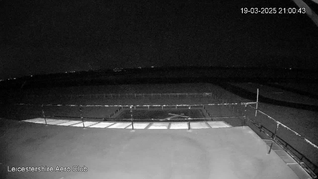 A dark image showing an aerial view of a runway and surrounding area. The foreground includes a section of a building roof with several lighted windows along the edge. A safety railing is visible on the right. In the distance, faint lights indicate the presence of other structures or roads. The overall scene is predominantly shadowy, suggesting it is nighttime. The timestamp indicates the image was captured at 21:00:43 on March 19, 2025.
