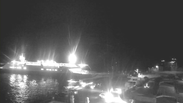 A nighttime image of a harbor scene. A brightly lit ferry is docked on the left, casting reflections on the water's surface. Several smaller boats are present near the dock, illuminated by lights. The surrounding area shows faint outlines of buildings and additional lights in the background, contributing to the nighttime ambiance.