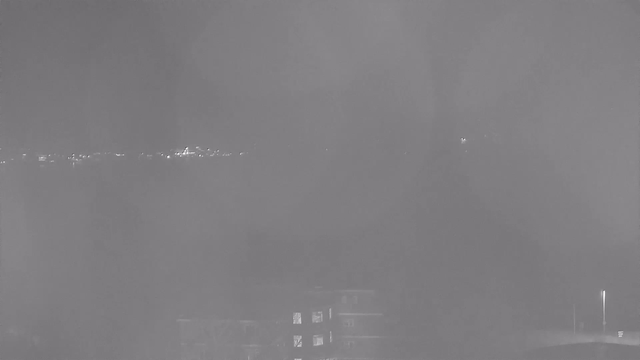 A dark, grainy night scene viewed from a webcam, showing faint outlines of a body of water and distant lights reflecting off the water's surface. In the foreground, shadowy shapes of buildings are visible, with a few illuminated windows. The overall atmosphere appears hazy and lacks clarity.