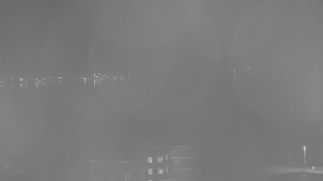 A dimly lit scene captures a view over water, with faint reflections of lights along the shoreline. The foreground contains an indistinct building outline with some illuminated windows. The overall atmosphere is hazy, suggesting mist or fog, which obscures details in the background.