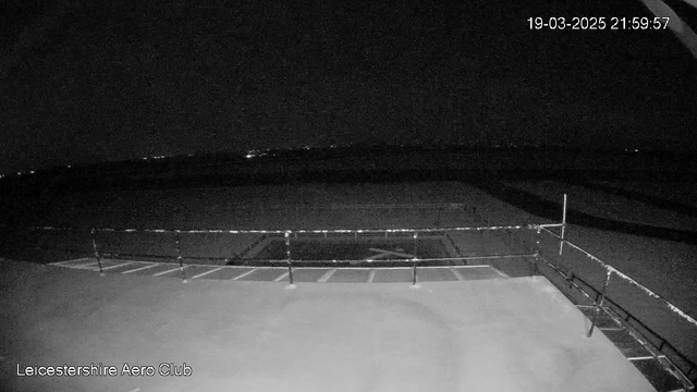A dark night scene from a webcam at the Leicestershire Aero Club. The foreground features a flat, concrete surface with markings for parking or maneuvering. In the background, there are dimly lit outlines of distant hills or structures, indicating a rural area. The date and time stamp reads March 19, 2025, at 21:59:57. The overall atmosphere is quiet and serene, typical of nighttime at an airfield.
