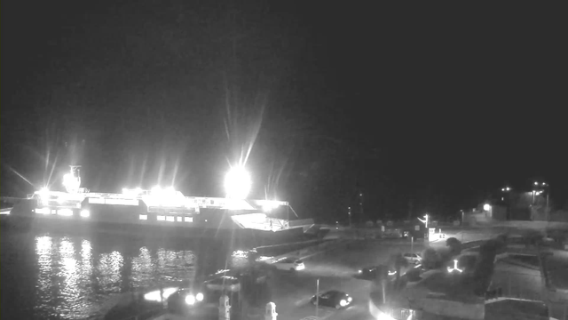 A brightly lit ferry is docked at a harbor at night. The ferry's lights reflect off the water, creating shimmering patterns. In the foreground, there are a few parked cars and a road. Several small lights from buildings and structures are visible in the background, indicating the presence of a town or port area. The overall scene is calm and predominantly dark, with the ferry being the main focus due to its illumination.
