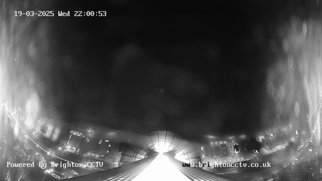 A black and white image from a webcam showing a night scene with a faint illuminated path leading towards the center. Surrounding the path are blurred lights indicating buildings and vehicles in the background. The date and time are displayed at the top of the image, along with a watermark at the bottom indicating it is powered by Brighton CCTV. The scene appears slightly hazy, possibly due to rain or mist.