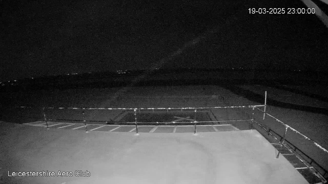 A dark, nighttime image captured from a webcam overlooking an open area with some structures. In the foreground, there are horizontal lines indicating a railing, and the surface below appears flat. In the background, faint lights are visible in the distance, suggesting the presence of a city or town. The sky is mostly dark with minimal illumination. A timestamp in the upper right corner indicates the date and time of the capture.