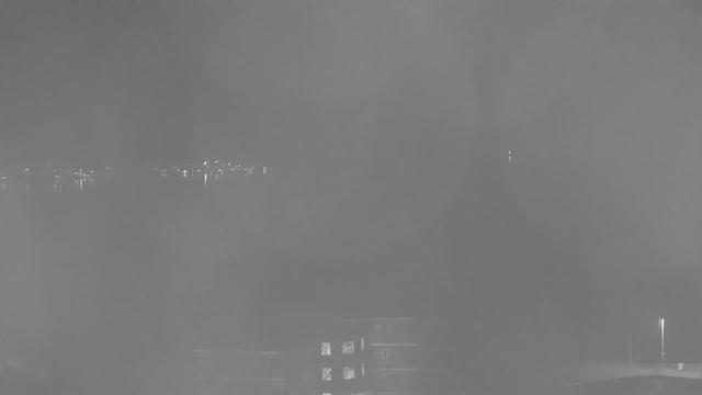 A gray-scale image shows a foggy or misty scene with limited visibility. In the distance, faint lights are visible reflecting on water, suggesting the presence of buildings or structures along the shore. The forefront features a slightly illuminated building with several windows, while the background is almost blank due to the haze, creating a soft and indistinct atmosphere.