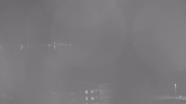 A dark and foggy scene showing a body of water with faint reflections of lights from a distant shoreline. A building is visible in the foreground, partially obscured by mist. The overall atmosphere is dim and unclear due to the fog.