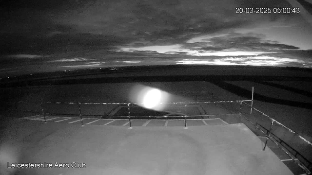 A dimly lit view from a webcam showing an outdoor area. The foreground features a flat surface with a railing, while the background displays a dark sky filled with clouds. The image is taken before dawn, with minimal visibility and a light glow near the left side indicating a distant light source or reflection.