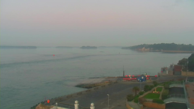 A tranquil waterfront scene at dawn, featuring calm waters that reflect a soft pink and blue sky. In the distance, a series of small islands are partially obscured by mist. Closer to the foreground, the shoreline reveals a harbor with a few boats. On the left, a red buoy is visible in the water, while the right side showcases a paved area with neatly trimmed grass and small trees. The overall atmosphere is serene and quiet, suggesting early morning light.