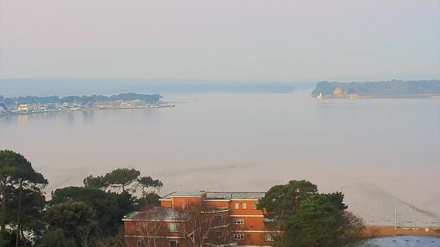 A serene water landscape at dawn, featuring a calm body of water that reflects soft hues of light. In the foreground, a reddish-brick building with several windows is visible, surrounded by trees. To the left, a waterfront area with various structures and boats can be seen along the shore. In the distance, a small island with what appears to be buildings is present, framed by a hazy atmosphere. The overall scene conveys tranquility and natural beauty.
