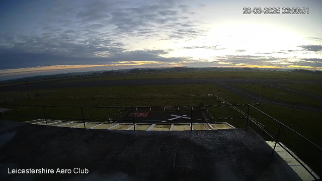 A wide view of an aerodrome at dawn, featuring a vast expanse of grassy land with a runway visible in the foreground. The sky is partly cloudy, transitioning from dark to light as the sun rises. The horizon shows varying shades of orange and gold. In the bottom left corner, there is a partial view of a rooftop railing and a section of the building. Markings on the runway include white lines and a large white 'X' on a red background, indicating a designated area.