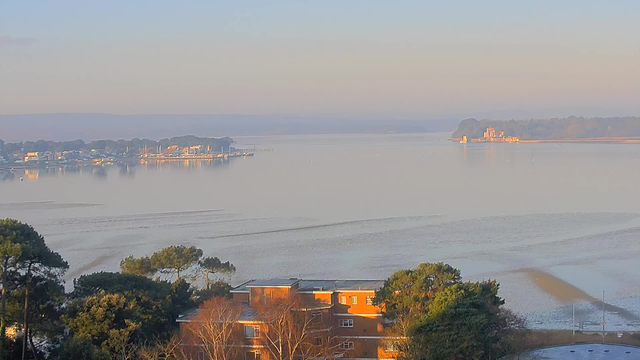 A tranquil view of a calm waterway during early morning. Soft pastel colors fill the sky, reflecting on the water's surface, which appears smooth and slightly rippled. In the distance, a small town is visible along the shoreline, with various buildings and boats docked. Silhouettes of trees frame the foreground, providing contrast to the serene scene. A distant land mass features a structure, possibly a building or fort, adding interest to the horizon. The overall atmosphere is peaceful and quiet, evoking a sense of calm.
