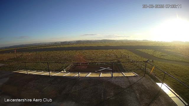 A clear blue sky stretches above a grassy airfield, with the sun rising in the background, casting a warm glow. In the foreground, there is a helipad marked with a white X. The ground is mostly flat with a few distant rolling hills visible on the horizon. A winding road runs across the airfield, and there are some wooden posts lining the area. The image captures a serene morning scene at the Leicestershire Aero Club.