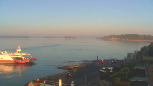 A tranquil scene of a harbor at dawn. In the foreground, a white and orange ferry is docked. The calm water reflects soft morning light, with a light haze over the horizon. There are a few small boats and a red buoy visible in the water. On land, a road runs beside the harbor, with parked vehicles and some structures, including a blue building and a small garden area. In the background, low hills are partially obscured by mist, suggesting a peaceful and quiet atmosphere.
