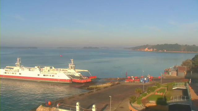 A ferry is docked at a port, with its ramp extended towards the land. The water is calm and reflects a clear blue sky, with a few distant green islands visible in the background. The shore features scattered trees and a small building, while red safety barriers are positioned along the dock. The scene is bright and quiet, suggesting a peaceful coastal environment.