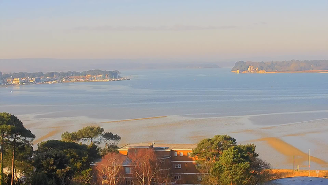 A panoramic view of a calm estuary surrounded by trees. The foreground features a few low-rise buildings with some greenery. In the distance, a small harbor is visible with boats, and a rocky coastline curves around the water. The sky is clear with a soft, early morning light illuminating the scene, creating a tranquil atmosphere.