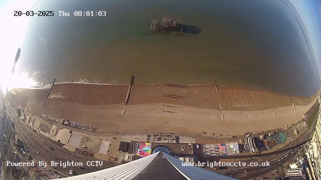 A view from a high vantage point overlooking a beach. The sandy shoreline stretches out with gentle waves meeting the water. A pier structure is visible a short distance from the shore, partially submerged. There are some people walking along the beach and a collection of buildings and attractions visible close to the shore, including colorful umbrellas and a large circular structure. The sky is bright, reflecting the sunlight over the water. The time and date are displayed in the top corner.
