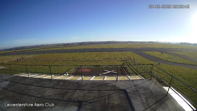 A clear blue sky is overhead, with some wispy clouds in the distance. Below, a grassy airfield stretches out, with a paved runway visible. In the foreground, there is a railing on a building balcony, and a helipad marked with a large white "X" on the ground. The scene is serene and open, suggesting a peaceful morning at the Leicestershire Aero Club.