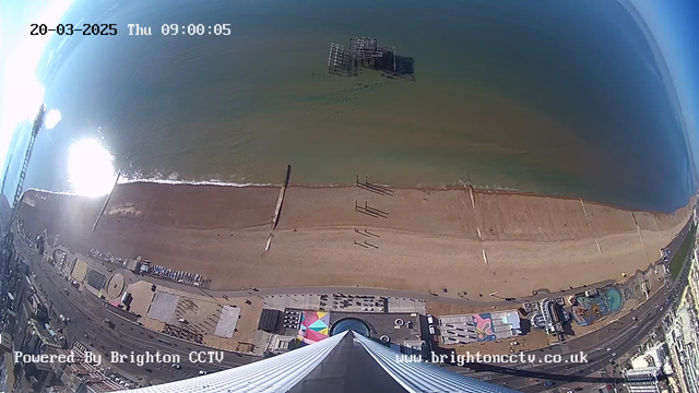 A high-angle view of a beach scene, with the ocean meeting the shore. The sandy beach features several people walking along the water's edge, with a wooden pier extending into the sea. In the background, there are some structures visible just offshore. The beach area is bordered by a promenade with various installations, including colorful patterns on the ground and amusement rides. The sky is clear, and sunlight reflects off the water, creating a bright glare. The image is timestamped and shows a date of March 20, 2025, at 09:00.