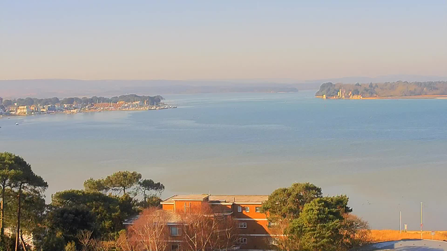 A serene landscape featuring a calm body of water with a pastel sky. In the distance, there are small boats and a marina lined with buildings along the shore. A tree-lined area frames the lower portion of the image, while gentle hills are visible in the background. The overall scene conveys tranquility and natural beauty.