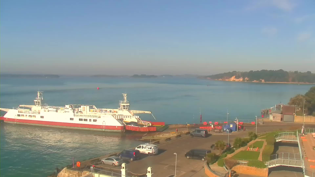 A calm waterfront scene featuring a large white and red ferry docked near a harbor. In the background, the blue water extends towards distant islands under a clear sky. The layout includes a parking area with several vehicles and some light greenery and pathways along the shoreline. A few small structures and a blue tent are visible near the edge of the harbor.