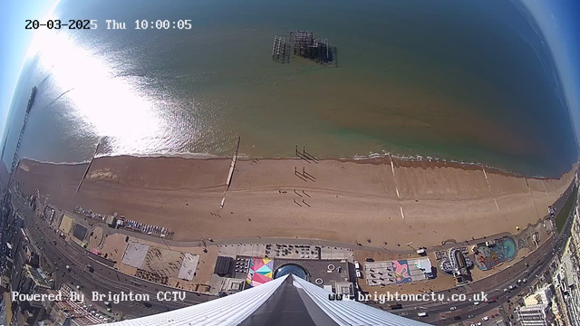 A bird's-eye view of a sandy beach along a coastline, with gentle waves lapping at the shore. In the distance, a pier is partially visible, and several people are walking along the beach. The sky is clear and blue, reflecting sunlight on the water, creating a shimmering effect. At the bottom of the image, there are colorful structures and a road with vehicles. The image is timestamped with the date and time at the top.