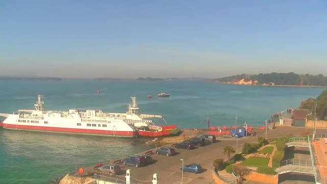 A ferry with a red and white hull is docked at a harbor, with a clear blue sky in the background. The ocean is calm, and a few small boats are visible in the water. In the foreground, there are parked vehicles along a paved area, with green grass and bushes lining the edges. Some buildings and a pier can be seen on the right side of the image, with various objects in the surrounding area.