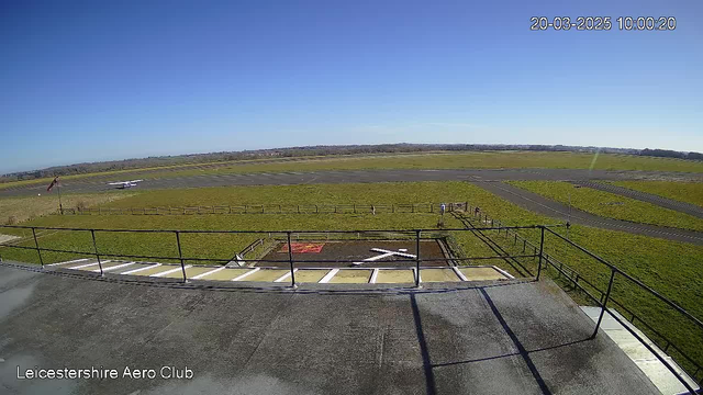 A view from the Leicestershire Aero Club's webcam shows a clear blue sky overhead. The grassy landscape includes an airstrip in the background, with some parked aircraft. A small group of people is seen walking near the edge of a fenced area. The scene is bright and open, indicating a sunny day.