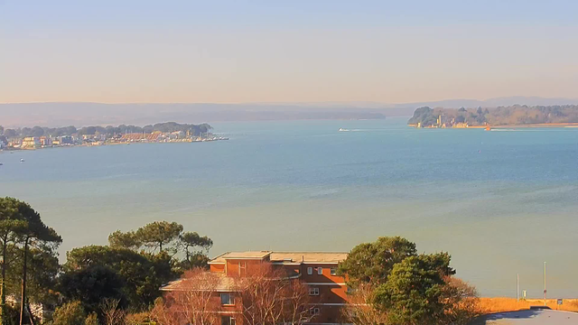A scenic view of a calm body of water, with soft blue and green hues. The shoreline features a marina with boats moored, and there are trees and buildings in the foreground. To the right, a small island with buildings is visible, along with a distant line of hills in the background. The lighting indicates a clear, sunny day, enhancing the tranquil atmosphere of the scene.