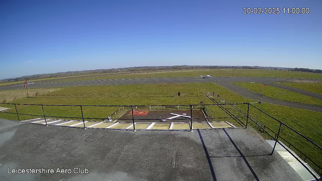 A clear blue sky fills the top half of the image, while the bottom portrays a view from a rooftop railing overlooking a grassy airfield. On the runway, a small airplane is taxiing. In the foreground, there is a weathered deck with a large white 'X' marking, and a few people and wooden fences are visible in the distance. The scene captures a serene day at the Leicestershire Aero Club.