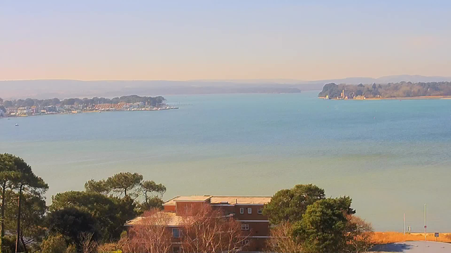 A scenic view of a calm bay with soft blue water. In the foreground, there are clusters of green trees. A low brown building is visible beneath the trees. Across the water, a shoreline features a variety of buildings and boats moored at docks. The background shows hazy mountains under a pale blue sky. The overall atmosphere is tranquil and serene.