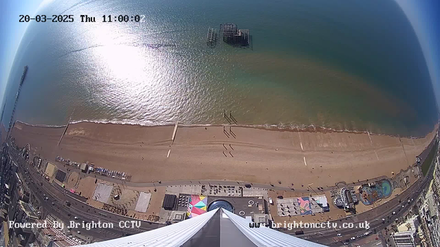 Aerial view of a beach on a sunny day. The foreground shows a sandy shoreline with a few people walking along the beach. The water is a mix of blue and green hues, reflecting sunlight. In the background, there is a wooden structure emerging from the water, likely a pier or ruined pier. Along the edge of the beach, colorful structures and rides suggest an amusement area. The sky is clear, indicating a bright day. The timestamp in the top corner reads 20-03-2025, 11:00:00.