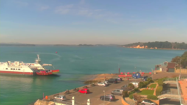 A waterfront scene featuring a ferry docked at a shore. The ferry has a red and white color scheme, with cars being loaded. The water is a calm turquoise, stretching towards the horizon where faint landforms are visible. The area is bordered by a parking lot with several parked cars, including red and white vehicles, and green grassy patches with small trees and shrubs. In the background, there are cliffs and greenery along the shoreline under a clear blue sky.