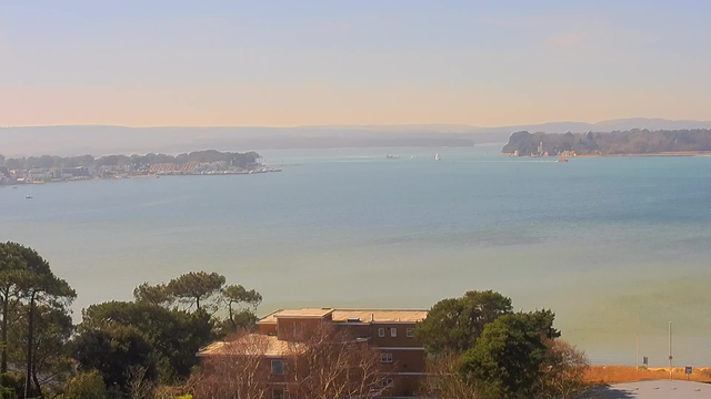 A serene waterfront view featuring calm blue waters under a clear sky. In the foreground, there are trees and a building with a reddish-brown exterior. To the left, a marina with several boats is visible, while the background showcases a distant shoreline and hills. The overall atmosphere is peaceful and bright, suggesting a sunny day.