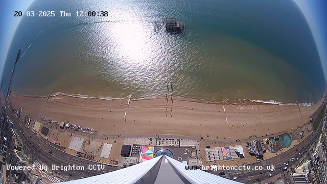 Aerial view of a beach on a sunny day, with gentle waves lapping at the shore. The sandy beach is dotted with people walking and a few scattered umbrellas. To the left, there is a pier extending into the water, while colorful beachside attractions, including a circular pool and structures, are visible near the bottom of the image. The ocean reflects sunlight, creating a shimmering effect. The sky is clear with no clouds.