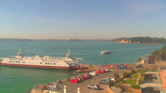 A coastal scene featuring a bright blue body of water under a clear sky. On the left, a large ferry boat is docked at a pier, while another smaller ferry is moving in the distance. In the foreground, a parking area filled with red cars is visible, alongside some green shrubbery and steps leading up to a walkway. The shoreline in the background is lined with trees and a sandy cliff.