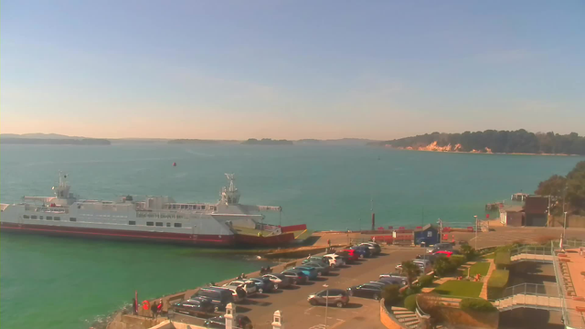 A serene waterfront scene featuring a large ferry docked at the edge, surrounded by calm blue waters. In the foreground, there is a parking area filled with various cars, with some people walking nearby. The background includes a distant shoreline with trees and sandy cliffs, under a clear sky.