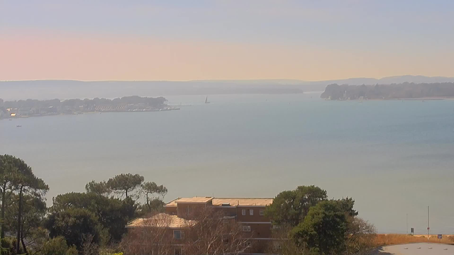A serene waterfront scene showing a light blue body of water with gentle waves. In the distance, a few small boats are visible near a shoreline dotted with trees and buildings. On the right side, there is a beach area with sparse vegetation and a road leading to the water. A light pink and blue sky creates a calm atmosphere, with distant hills under the horizon on the left.