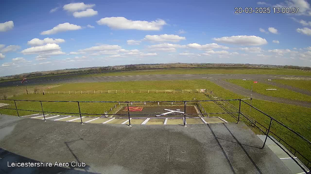 A view from a webcam at Leicestershire Aero Club shows a wide, open landscape under a clear blue sky with scattered white clouds. In the foreground, there is a flat roof with a railing surrounding it. Below, a grass field and a runway are visible, marked with white lines. A small red flag is present on the left side of the image, and a sign is visible near the edge of the grass area. The scene appears sunny, indicating a bright day.