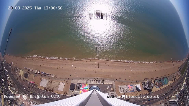 Aerial view of a beach and coastline. The beach has a sandy shore with some people walking. In the distance, waves are gently breaking along the water's edge. There's a pier extending into the water, and several colorful beach huts are visible along the shore. A row of buildings can be seen in the lower portion of the image, along with various structures and a circular area. The sky is clear with some visible sunlight reflecting off the water.
