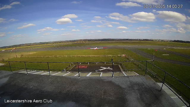 A wide view of an airport runway at Leicestershire Aero Club is depicted. The scene features a clear blue sky with a few white clouds. In the foreground, there is a railing visible, suggesting a balcony or observation area. The runway is visible in the distance, with a grassy area surrounding it. A small aircraft with a red and white design is parked on the runway. There are markings on the ground, including a large "X" symbol indicating a no-land zone.