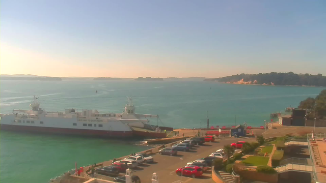 A coastal view featuring a blue sea and clear sky. In the foreground, a large ferry is docked at a terminal, with a few people visible near the vessel. To the right, there's a parking area filled with cars, including several red ones. In the background, green hills and rocky cliffs rise along the shoreline, while the water reflects the sunlight, creating a serene atmosphere.