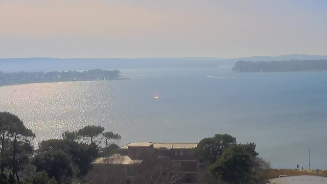 A tranquil view of a wide body of water under a clear sky. The sunlight sparkles on the water’s surface, creating a shimmering effect. In the foreground, there are green trees and a building with a flat roof, partially visible. In the distance, a landmass with trees is seen along the water's edge, and a small boat is navigating the water. The horizon shows gentle hills fading into the distance.