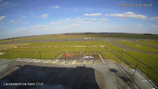 A view from a webcam at Leicestershire Aero Club. The scene features a grassy airfield under a clear blue sky with a few white clouds. In the foreground, there is a flat rooftop area with a railing and markings, including an "X" on the ground. A small aircraft is positioned on the runway in the background, with a long stretch of grass beside the runway and a wooden fence enclosing part of the space. The time displayed shows it is 3:00 PM on March 20, 2025.